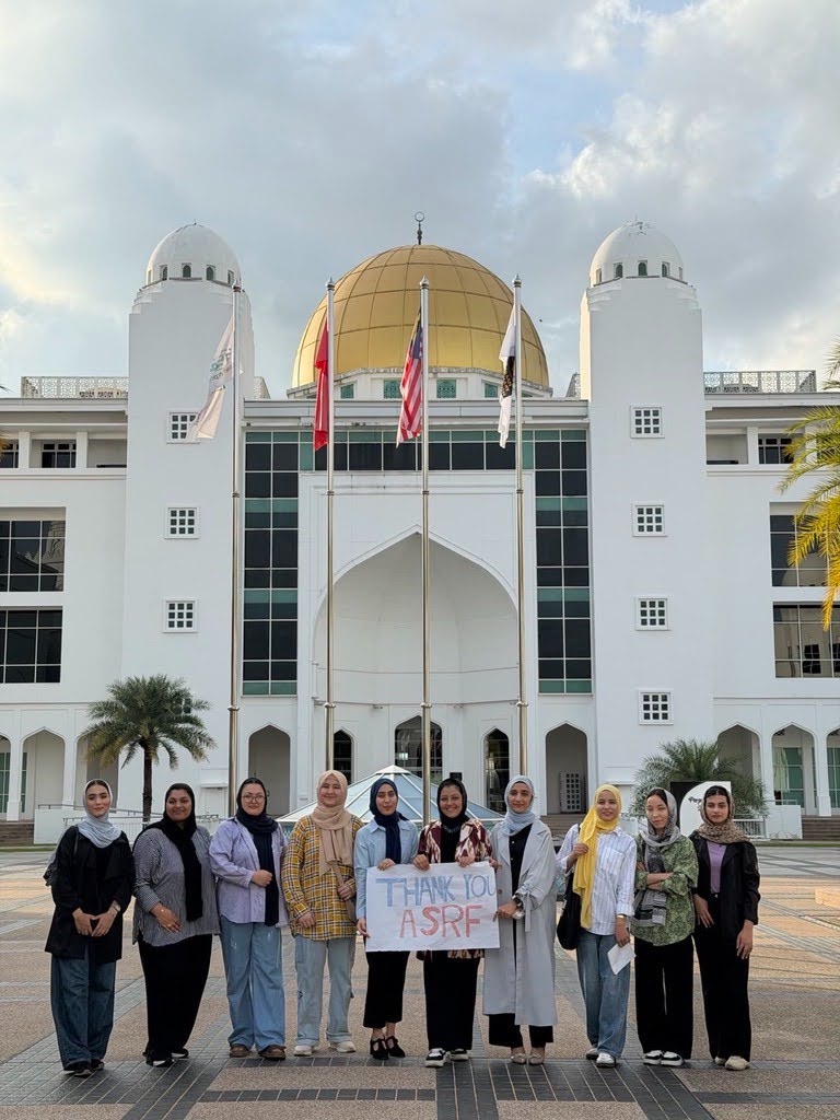 Nine Afghan women students standing in front of the white domed main building of Albukhary International University, holding a hand-lettered thank-you sign for ASRF