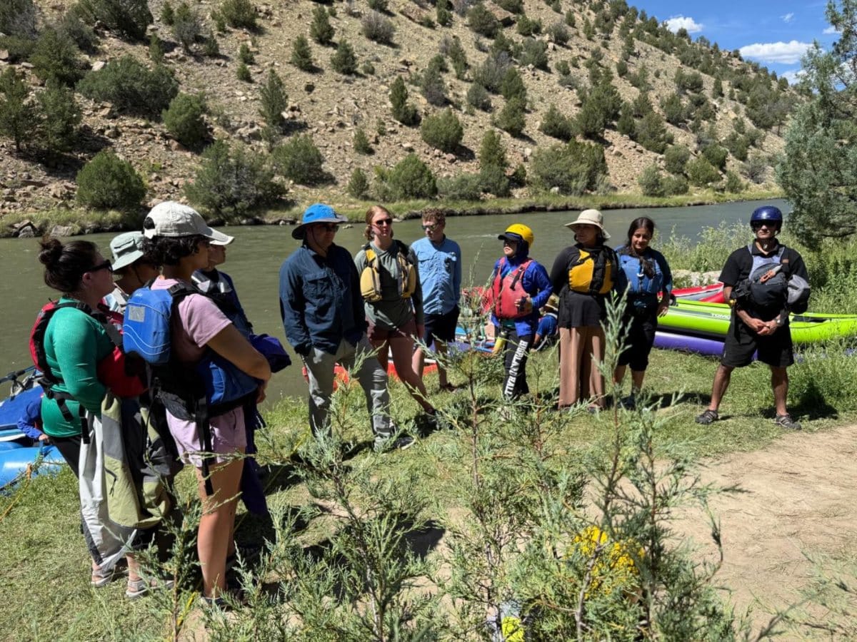 Scouts with backpacks gathered beside a river, kayaks pulled up on the bank behind them