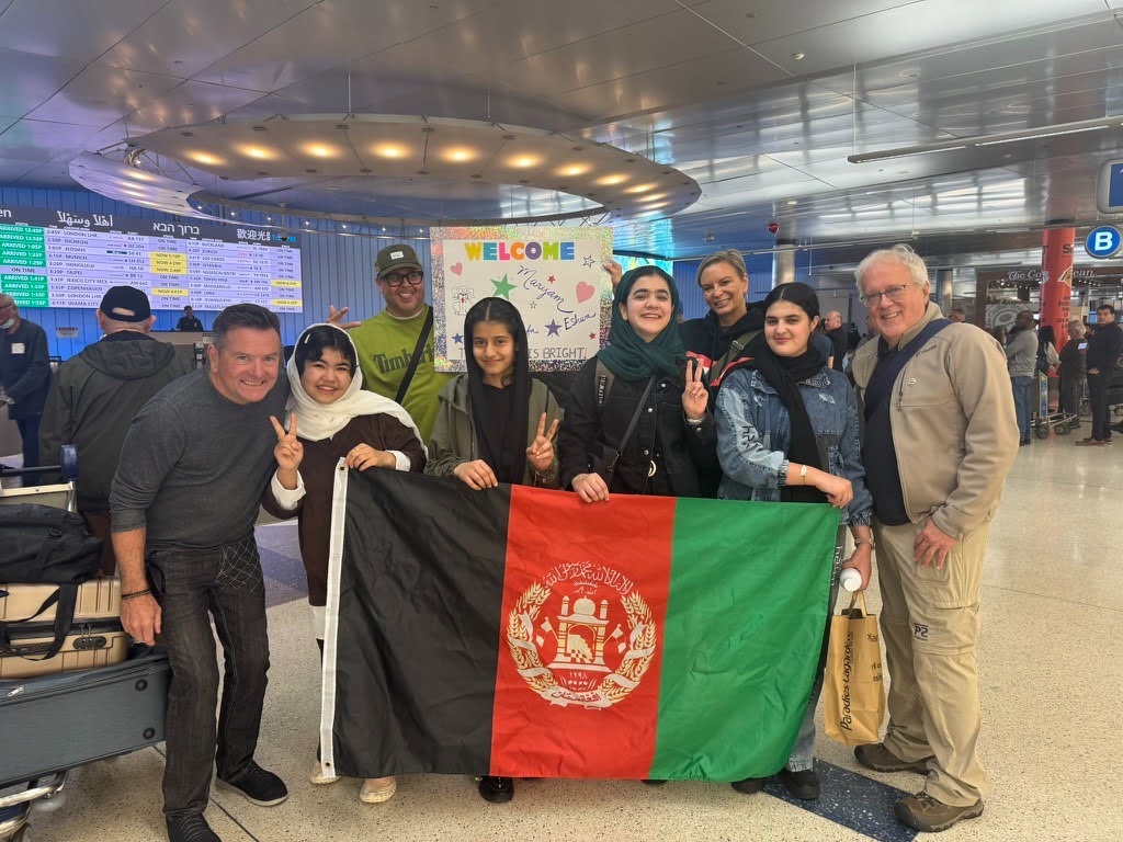 ASRF students and welcomers holding the Afghan flag beneath a hand-made welcome sign at the airport