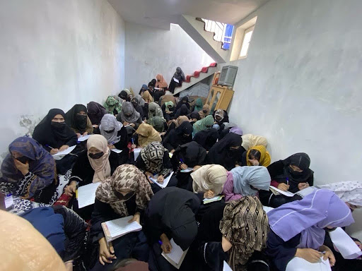 Dozens of Afghan women seated closely together, writing in notebooks in an underground classroom