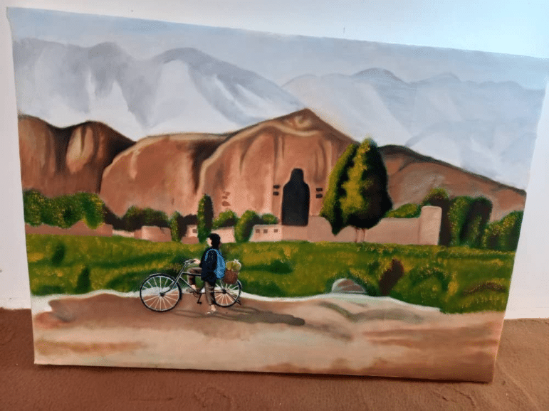 Painting of an Afghan village at the foot of cliff-face caves, with a person walking a bicycle along a dirt road