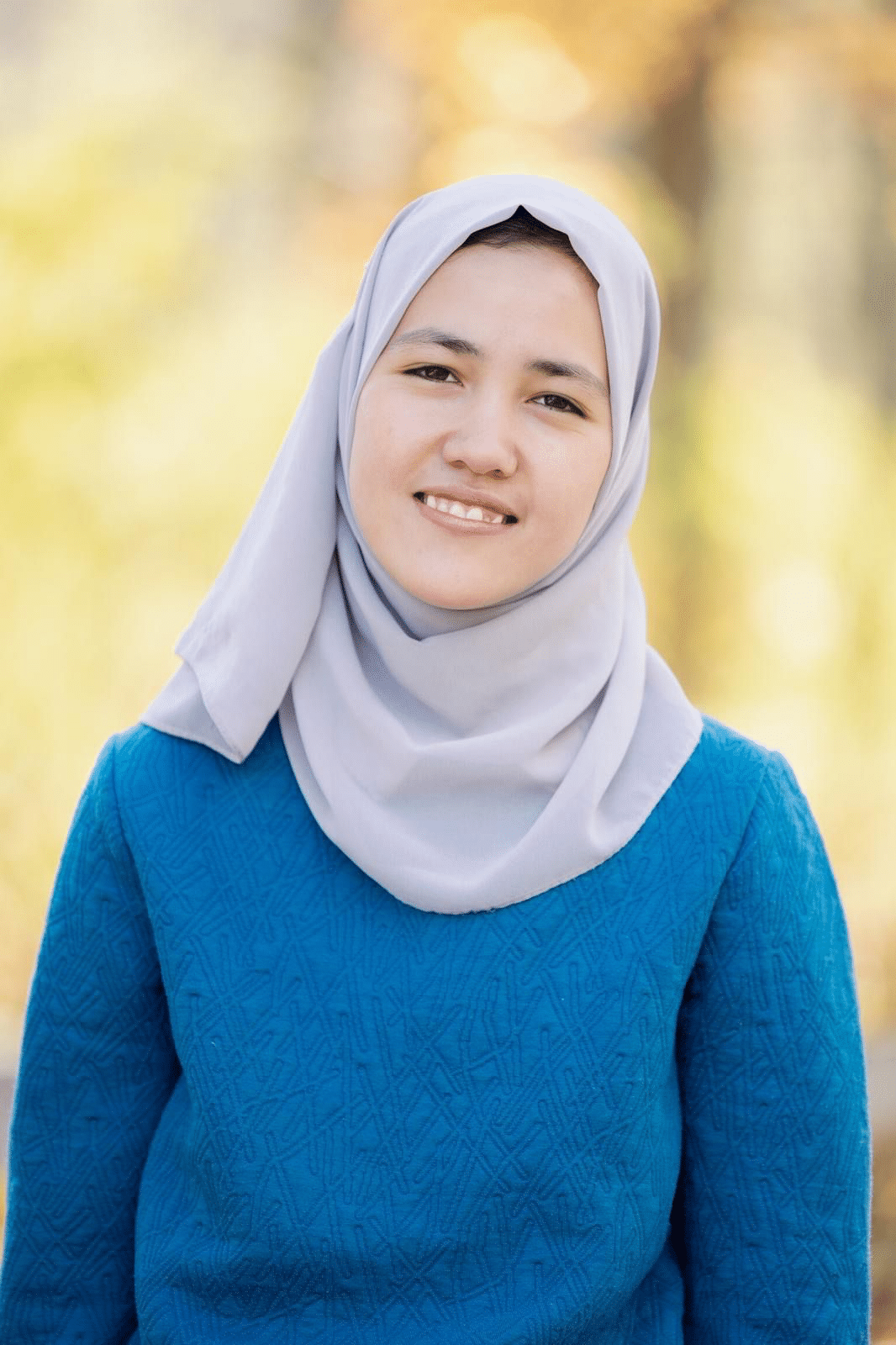 Masuda smiling outdoors, wearing a grey headscarf and blue sweater